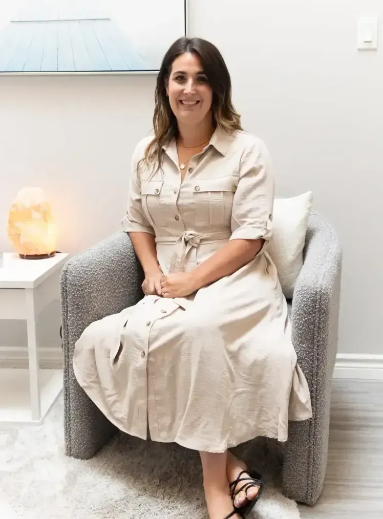 Woman sitting in armchair, smiling. Wearing a tan dress, black sandals, in a room with a salt lamp.