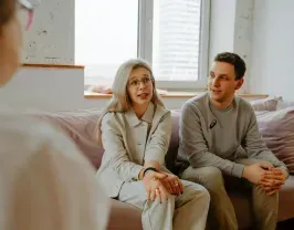 Couple in counseling session, on couch, speaking to a therapist. Light colors, natural light.