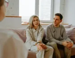 Couple in counseling session, on couch, speaking to a therapist. Light colors, natural light.