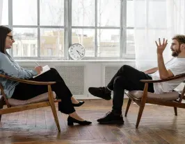 A therapist listens to a client. They sit in chairs facing each other near a window. The therapist takes notes.