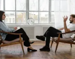 A therapist listens to a client. They sit in chairs facing each other near a window. The therapist takes notes.