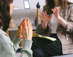 Two women in a therapy session; one gestures, the other clasps hands, with a notebook in front of them.