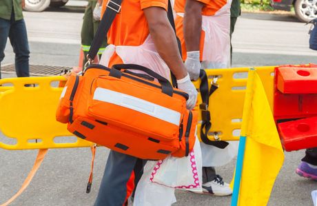 Paramedic with an orange medical bag, near a yellow stretcher, at an outdoor scene.