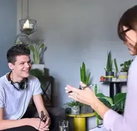 Two people chatting indoors, one smiling on a couch with headphones around his neck, plants in the background