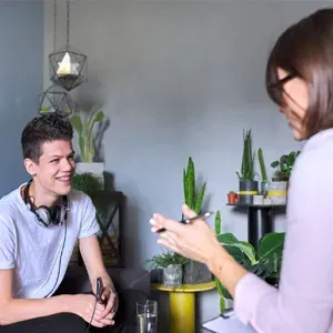 Two people talking in a plant-filled room, one seated and smiling while the other stands nearby holding a phone.