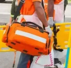 Person carrying an orange medical bag, near a stretcher and other emergency equipment.