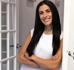 Woman with long dark hair smiles, arms crossed, leaning against a white doorway, bright setting.