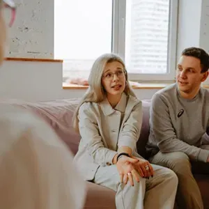 Two people sit on a couch in a bright room, talking with someone off-camera.