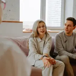 Two people sit on a couch in a bright room, talking with someone off-camera.