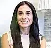 Smiling woman with long dark hair in a light pink top, seated indoors near a window.