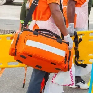 Person in orange safety vest carrying an orange emergency bag near a yellow barrier