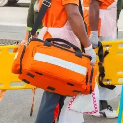 Person in orange safety vest carrying an orange emergency bag near a yellow barrier