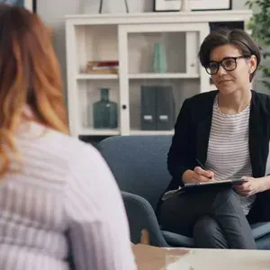 Two people in a counseling session, one taking notes on a clipboard in an office lounge.