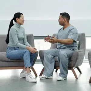 Two people seated and talking in a bright room, wearing casual clothes and sneakers.