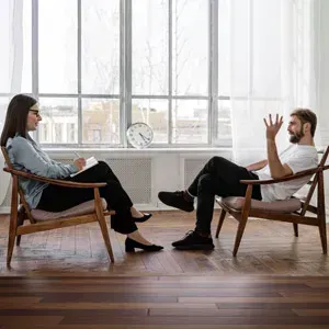 Two people seated in chairs talking across from each other in a bright room with large windows.