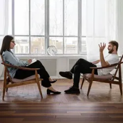 Two people seated in chairs talking across from each other in a bright room with large windows.