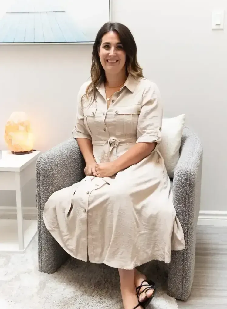 Woman sitting in armchair, smiling. Wearing a tan dress, black sandals, in a room with a salt lamp.