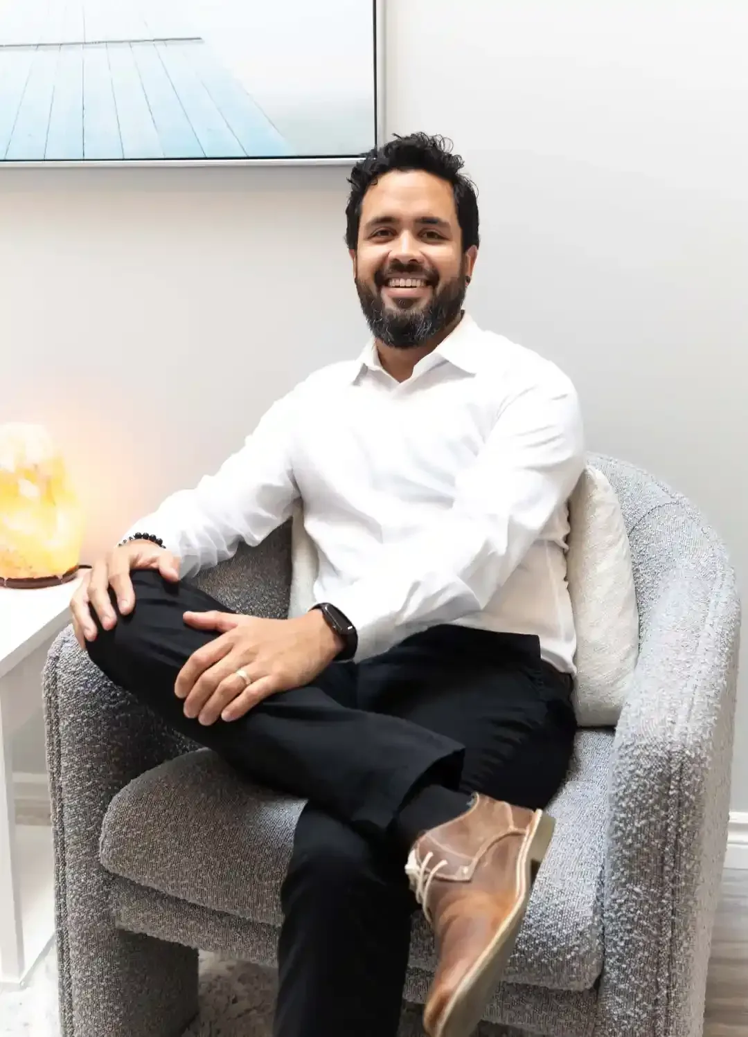 Man sitting in a chair, smiling, wearing a white shirt and black pants.