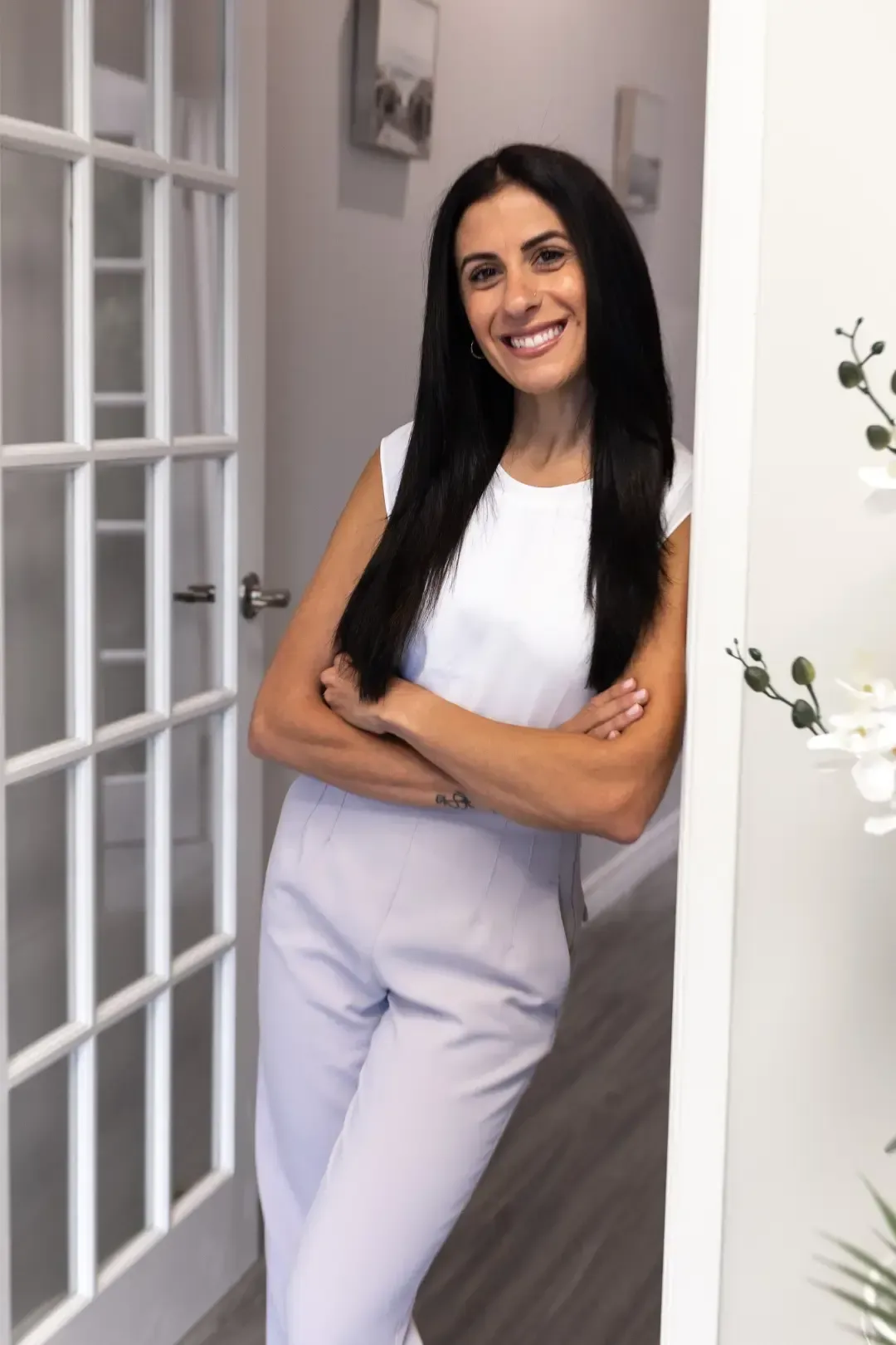 Woman smiling, arms crossed, leaning in doorway; white top, light pants.