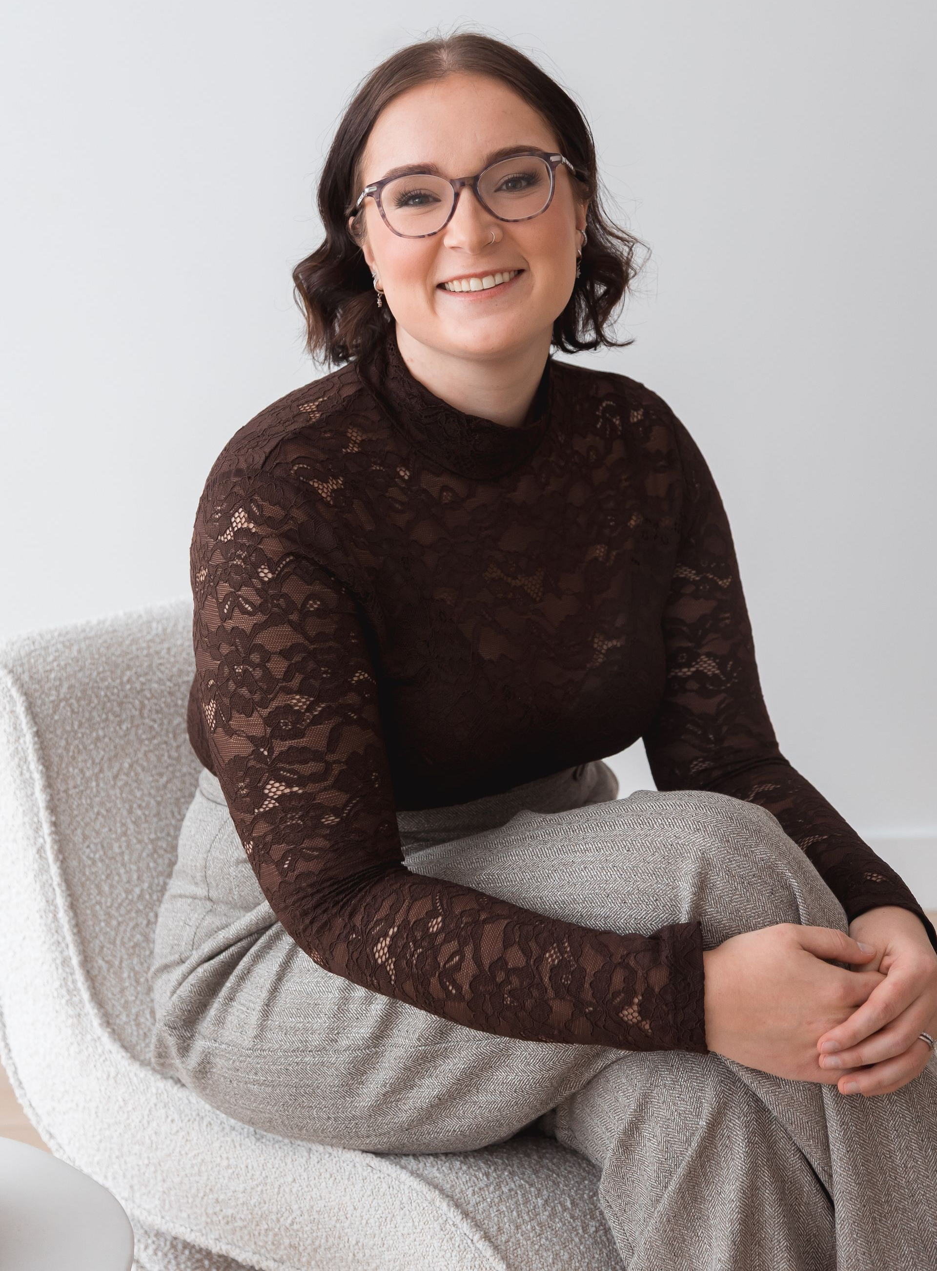 Woman with glasses smiles while sitting in a gray armchair, wearing a black top and jeans.