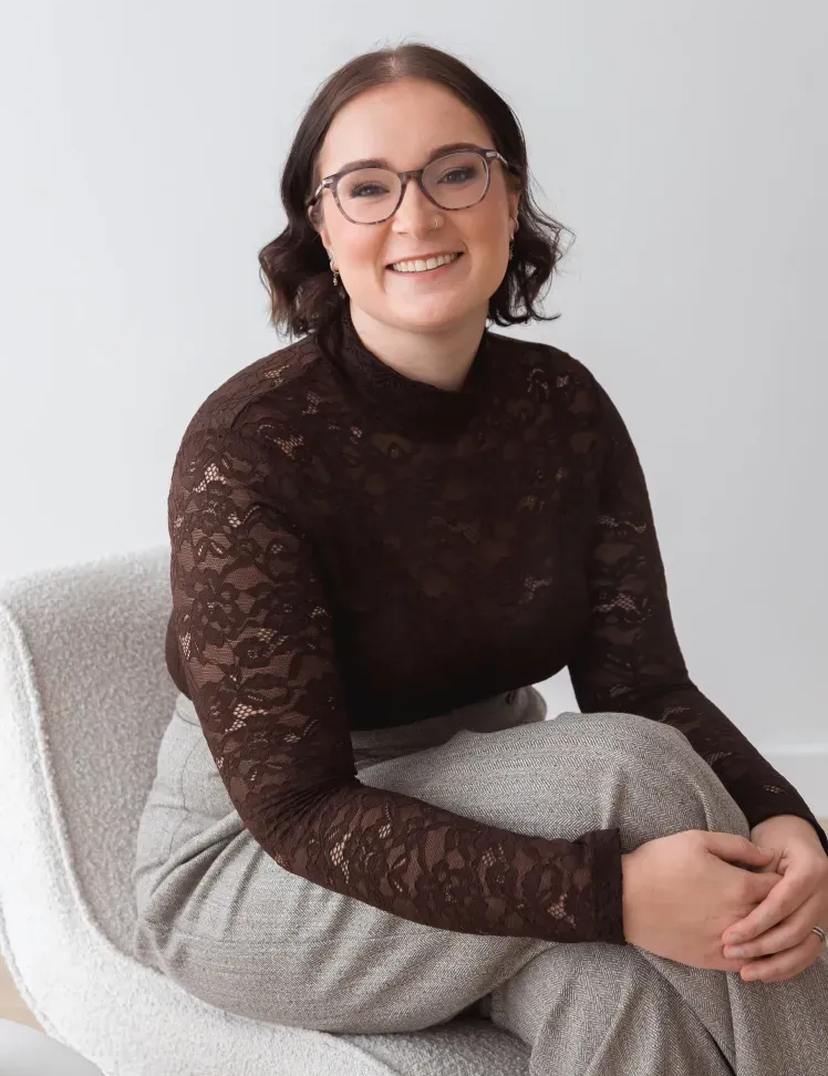 Woman with glasses smiles while sitting in a gray armchair, wearing a black top and jeans.