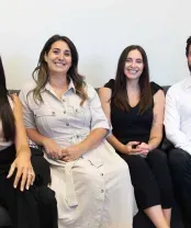 Four people smiling, seated close together on a gray couch, indoors.