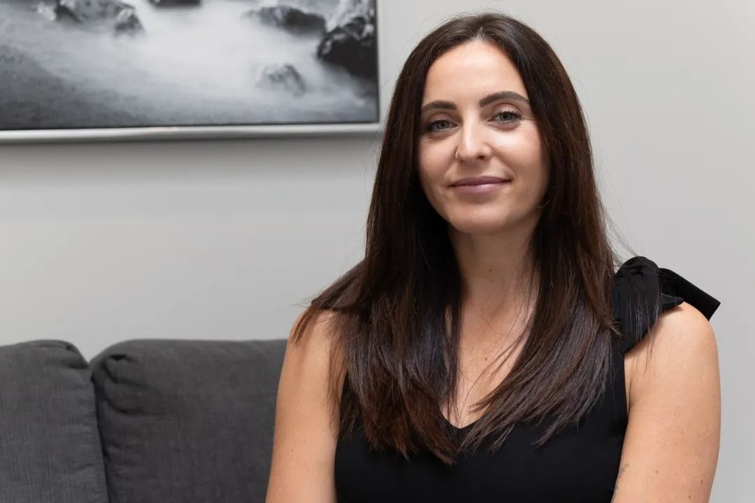 Woman with long dark hair, wearing a black top, sitting on a gray couch in front of a framed monochrome artwork.