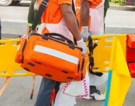 Person carrying an orange medical bag, near a stretcher and other emergency equipment.