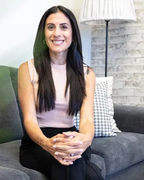 Woman with dark hair smiles, sitting on a gray couch in a room with a lamp and brick wall.