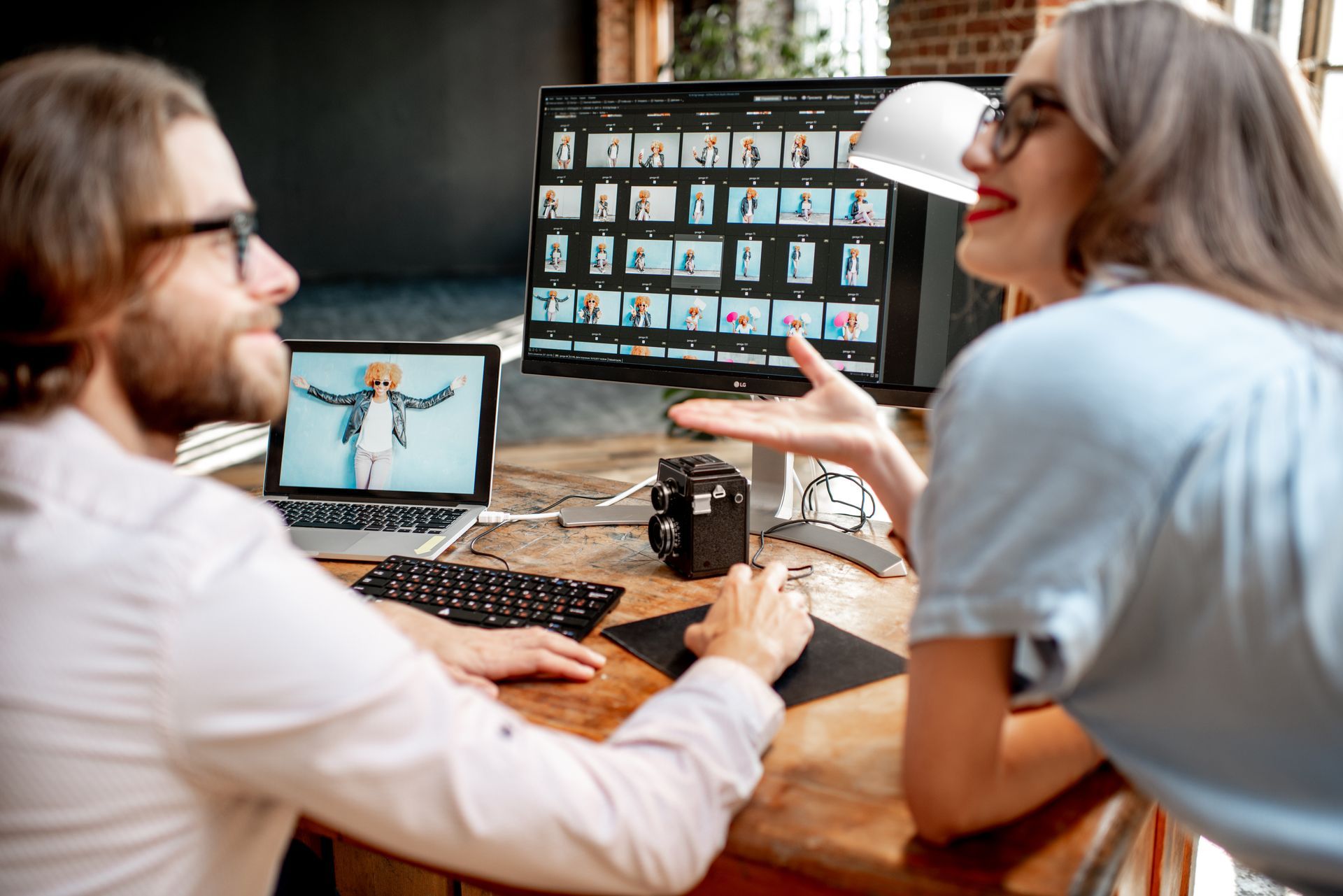 Two people reviewing photos on a computer and laptop, smiling and talking. A camera sits nearby on a wooden desk.