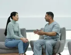 Woman in jeans and a sweater chats with a person in scrubs in a waiting room.