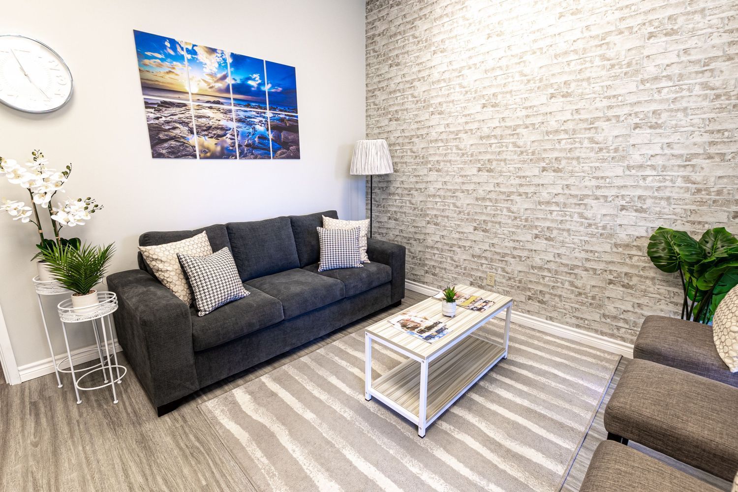Waiting room with gray sofa, brick wall, artwork, and a rug.