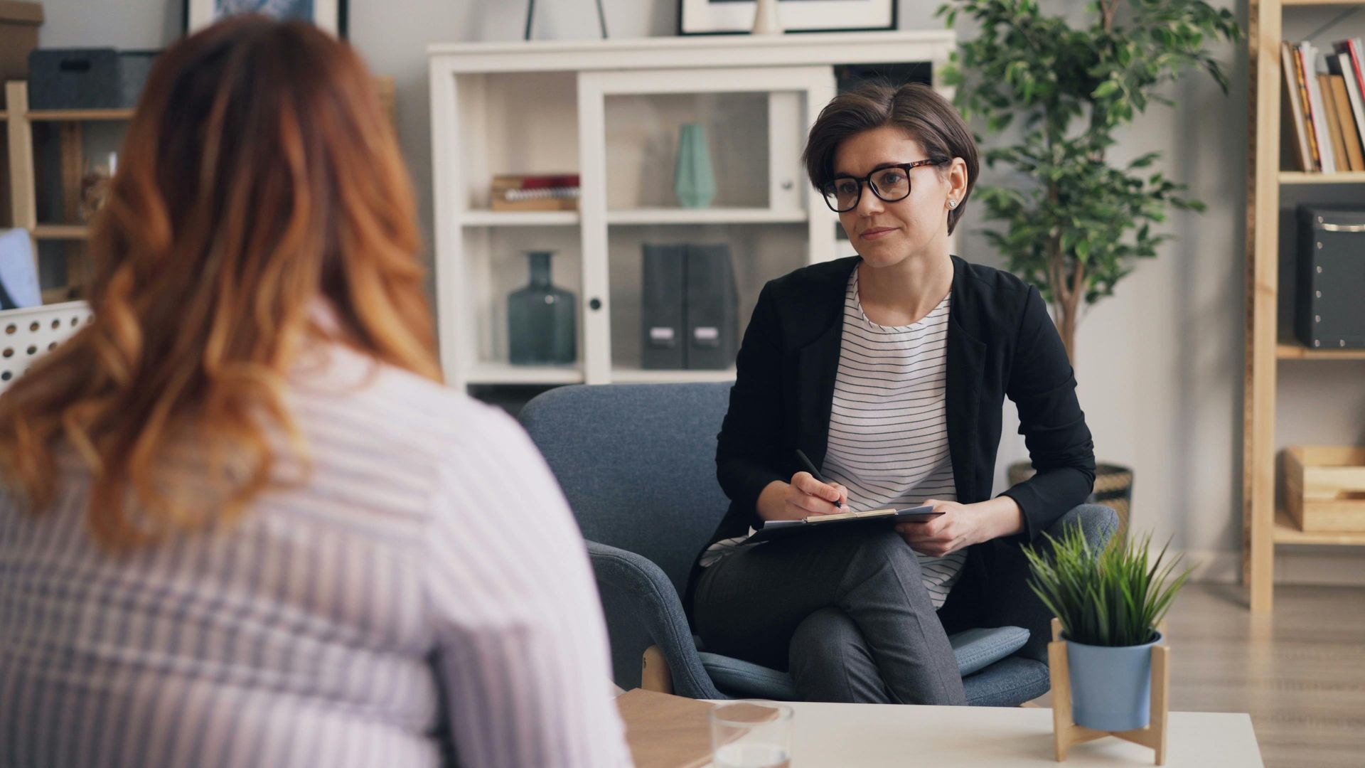 Therapist and patient in a counseling session. Woman with glasses takes notes; patient faces away.