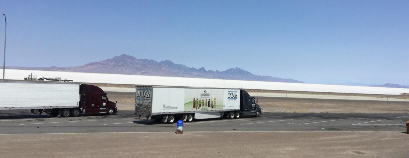 Two semi trucks are parked in a parking lot with mountains in the background.