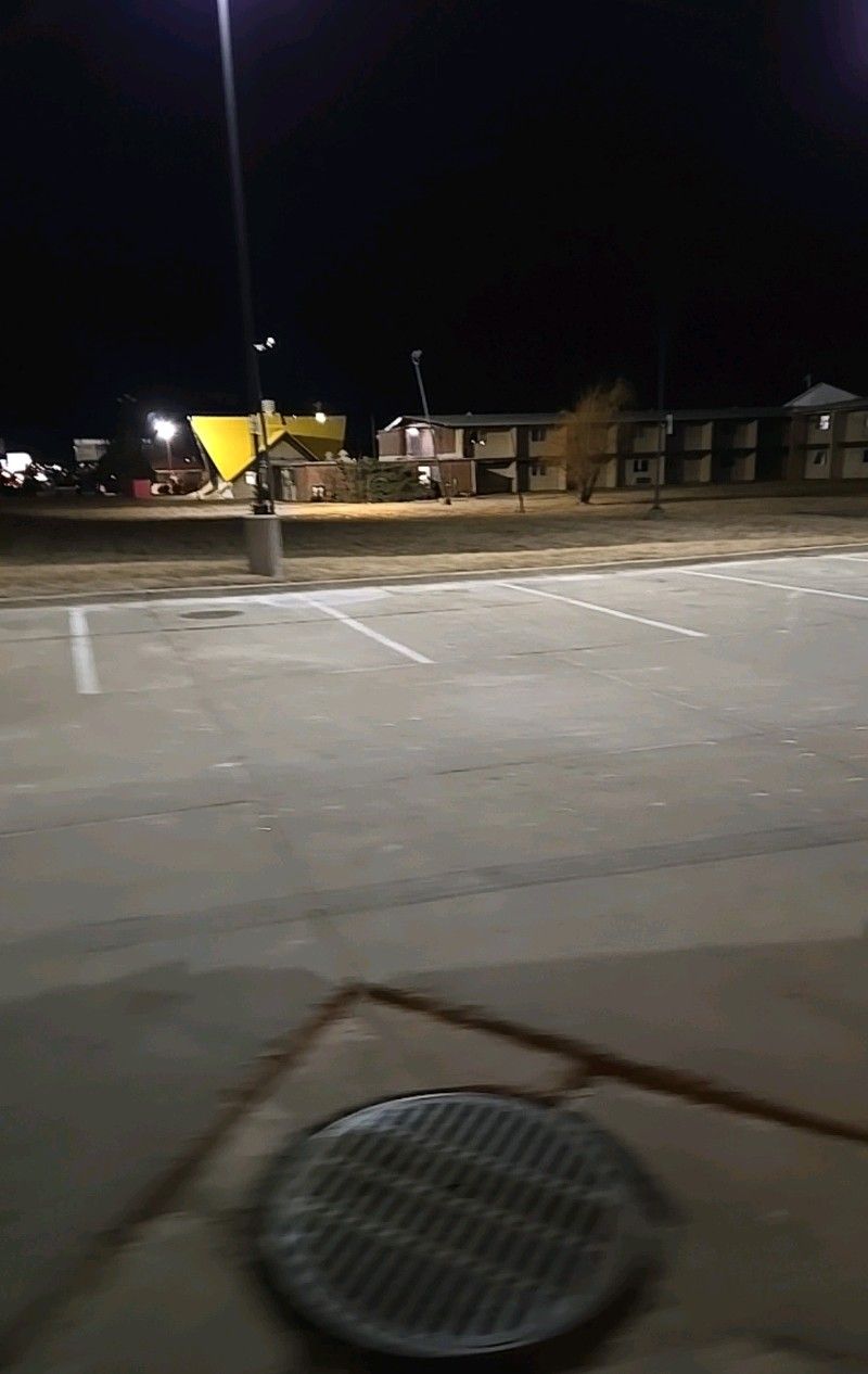Night shot of a nearly empty parking lot, illuminated by streetlights, with a building in the background.