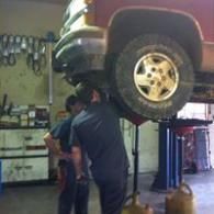 Two mechanics in a garage work on the undercarriage of a raised red vehicle, with spare parts hanging on the wall.