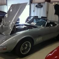 A silver vintage Chevrolet Corvette convertible with its hood open, parked inside a garage.