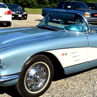 A vintage light blue and white Chevrolet Corvette convertible parked in a gravel lot with other vehicles.