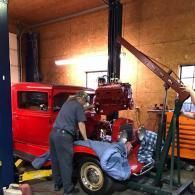 A mechanic uses a hoist to install a red engine into a classic red car inside a workshop.