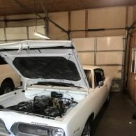 A white classic Plymouth Barracuda with its hood open, parked in a garage with a metal sectional door.