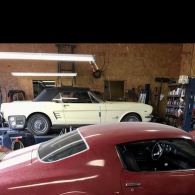A cream-colored convertible and a red muscle car parked inside a cluttered garage workshop.