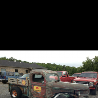 A rusted vintage pickup truck parked in a gravel lot next to several other vehicles and a brick building.