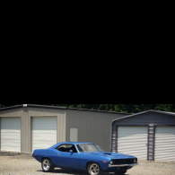 A blue classic muscle car parked in front of two metal garage buildings on a gravel lot.