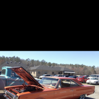 A vintage orange muscle car with its hood open sits in a parking lot on a sunny day.