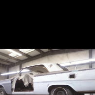 A light-colored vintage car body undergoing restoration in a well-lit workshop with overhead beams.