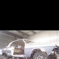 A primer-gray classic car body stripped to its frame, undergoing restoration inside a spacious, industrial workshop.