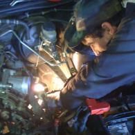 A person wearing a baseball cap and gloves repairs a car engine in a dimly lit garage using a work light.