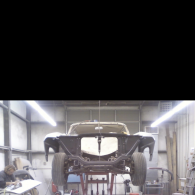 A car chassis is suspended on a lift in a workshop, with a person visible working at a nearby workbench.