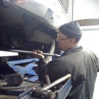 A mechanic wearing a beanie uses a long metal bar to work on the underside of a vehicle lifted in a garage.
