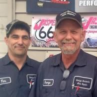 Two mechanics in navy work shirts stand in front of an automotive sign, smiling at the camera.